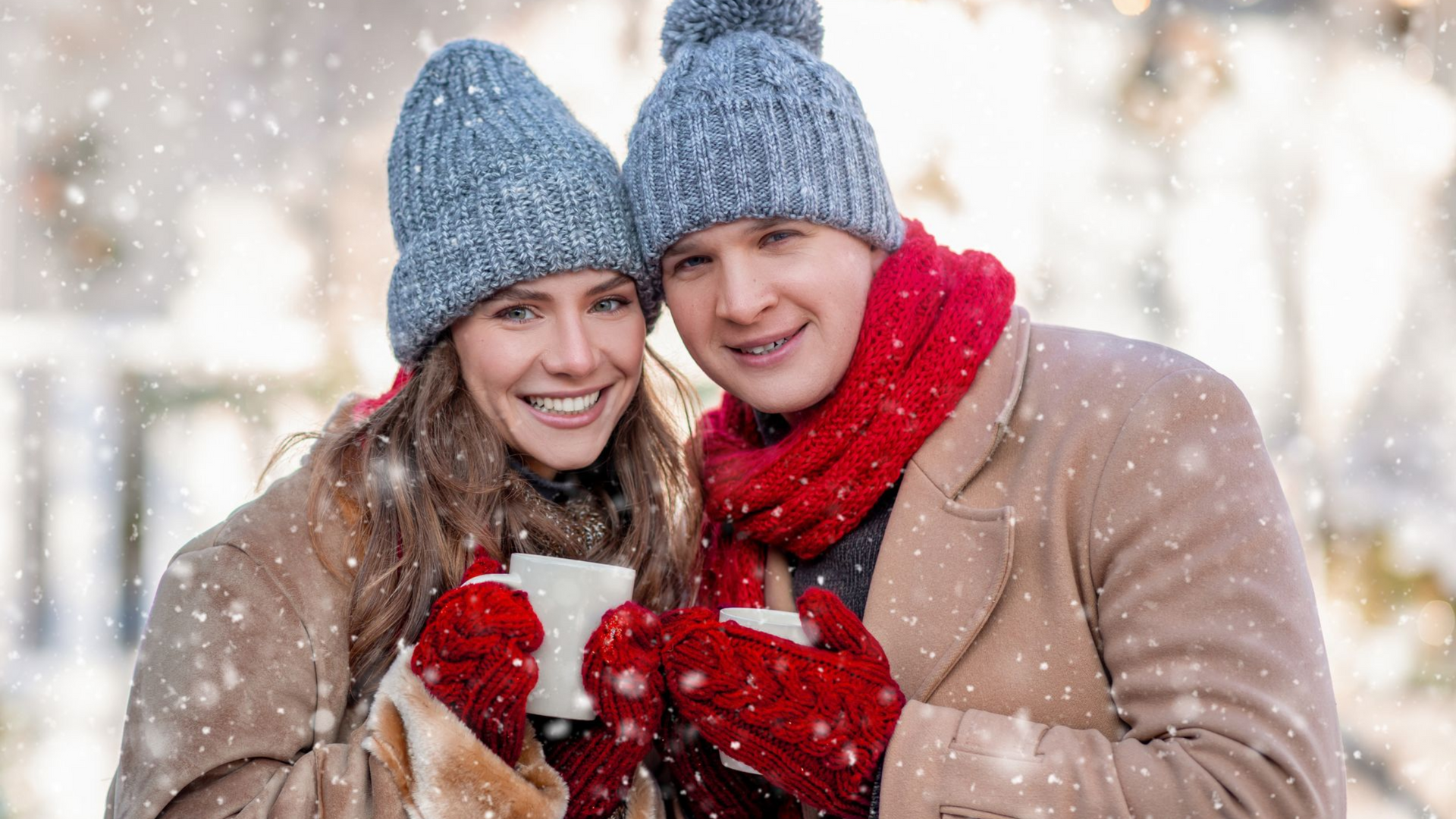 Couple in warm winter wear smiling, holding mugs outdoors in the snow.