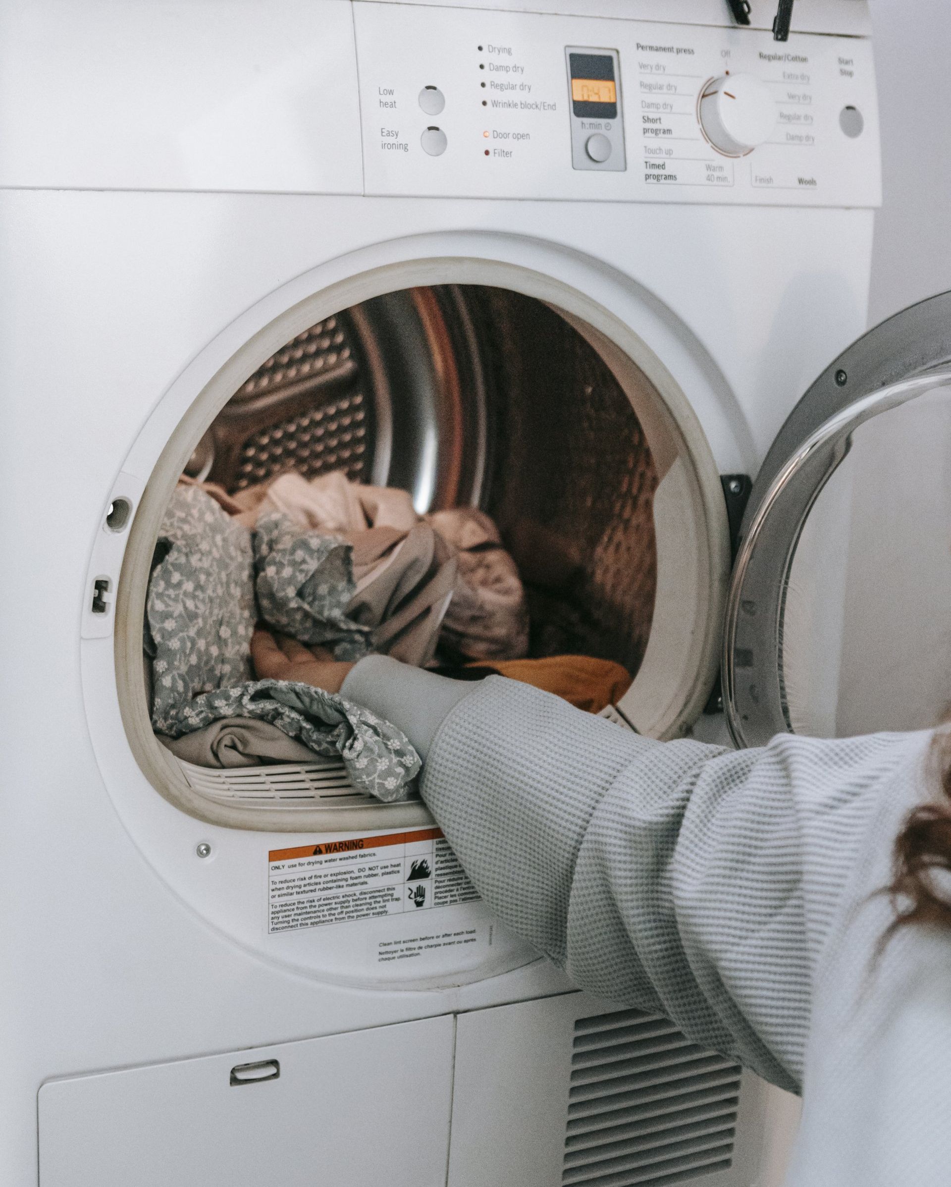 Hand placing a bundle of clothes into the open dryer drum, preparing for a fresh laundry cycle.