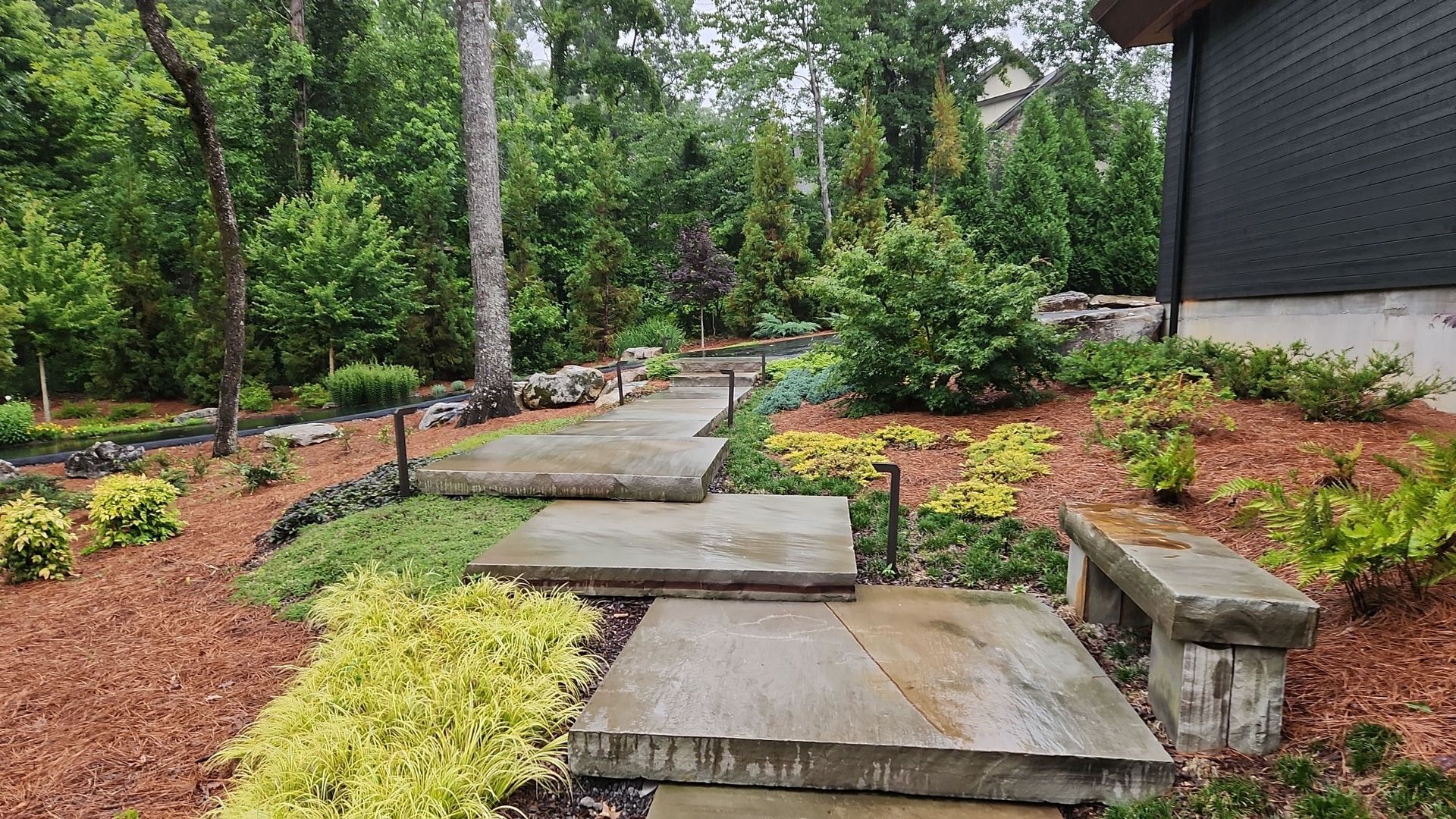 A stone walkway leading to a house surrounded by trees and bushes.