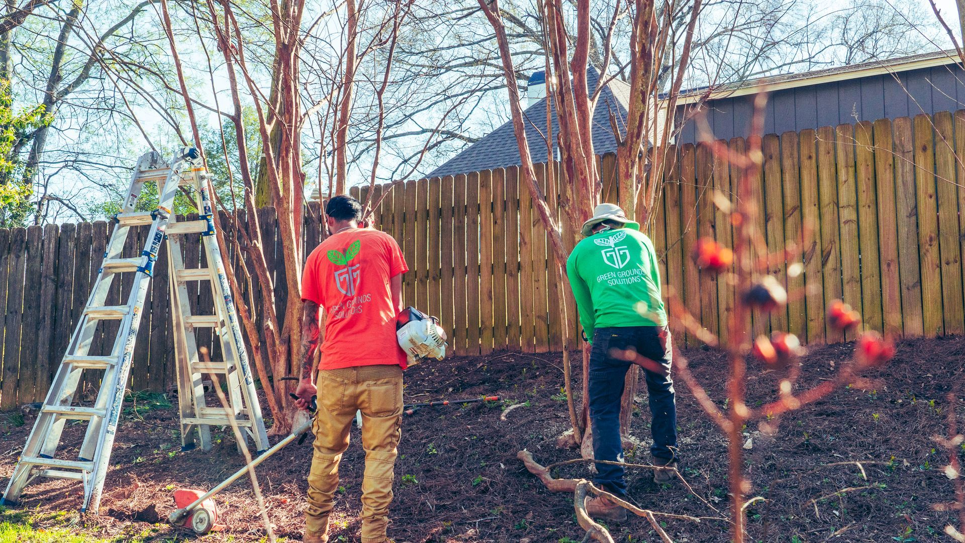 Two landscapers are standing in front of a wooden fence.
