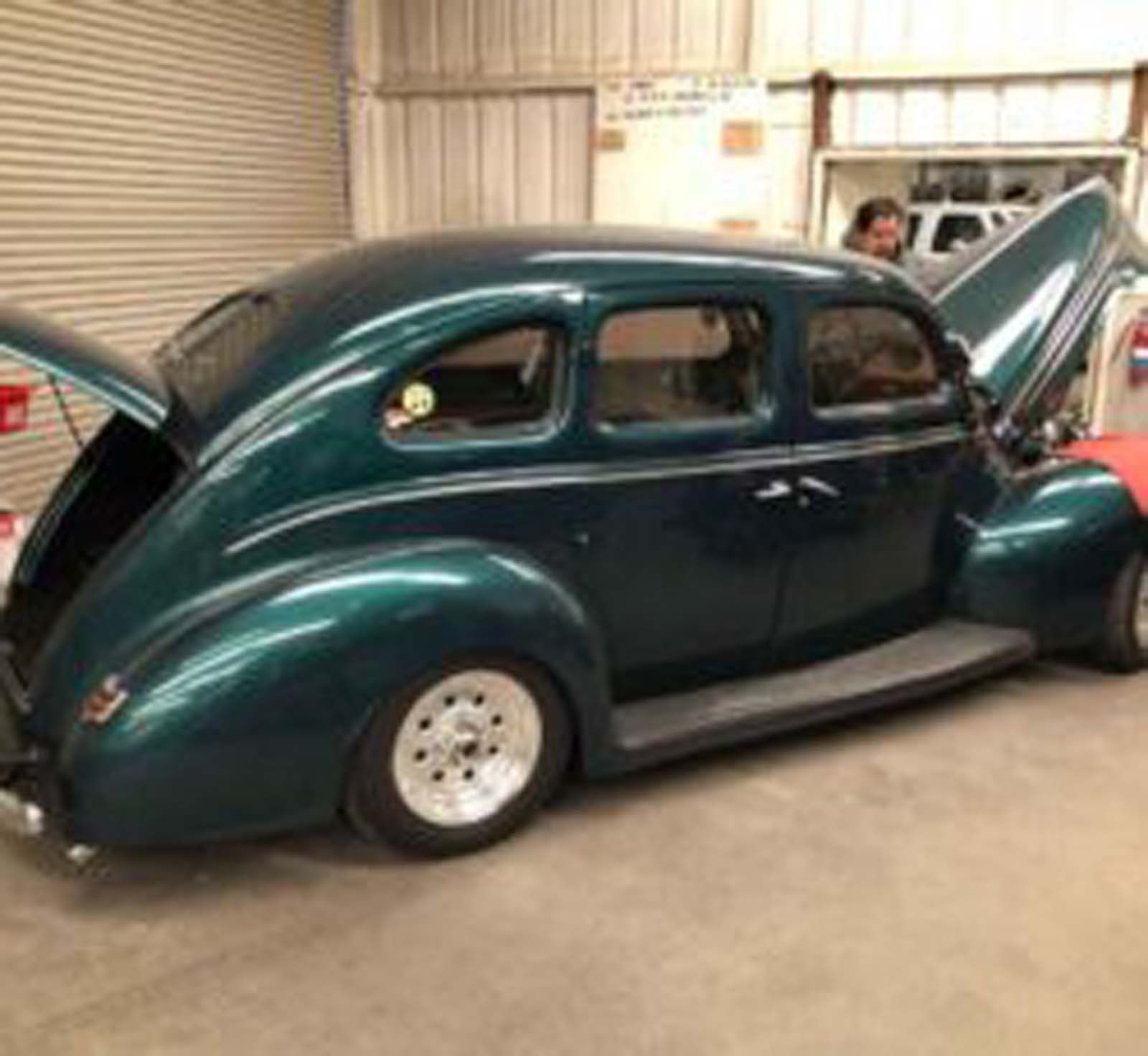 An old green car with the hood up is parked in a garage.