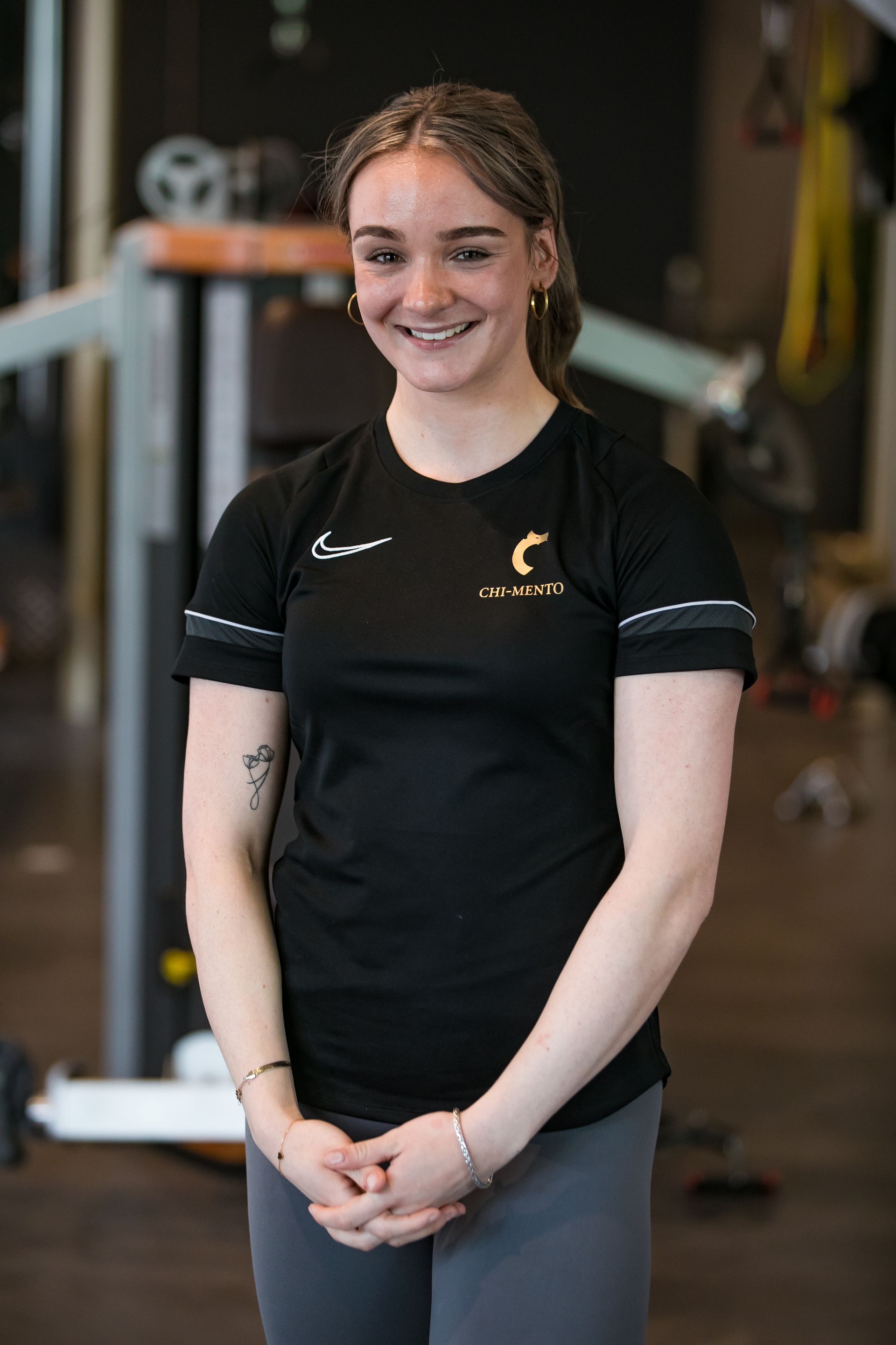 Een vrouw in een zwart shirt en grijze broek lacht in een sportschool.