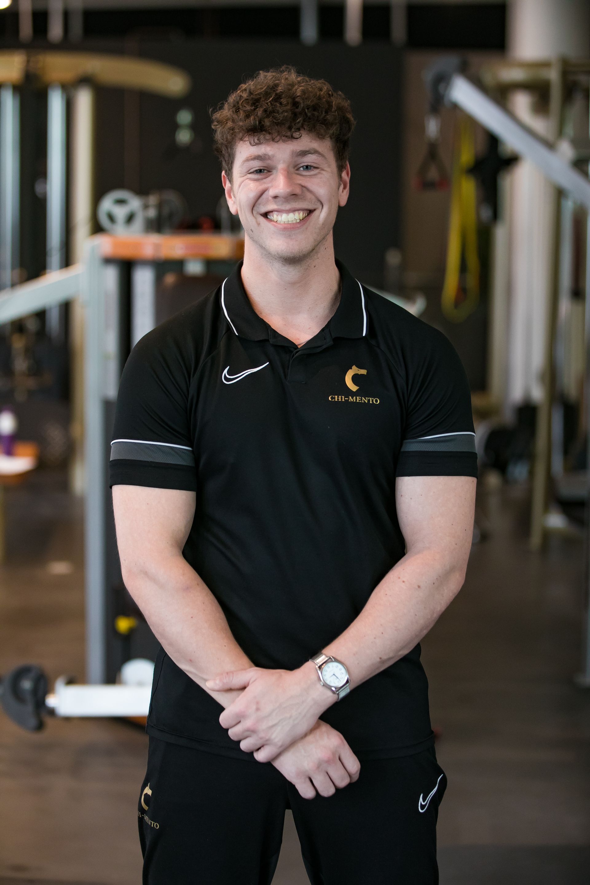 Een jonge man staat in een sportschool met zijn handen gevouwen en glimlachend.