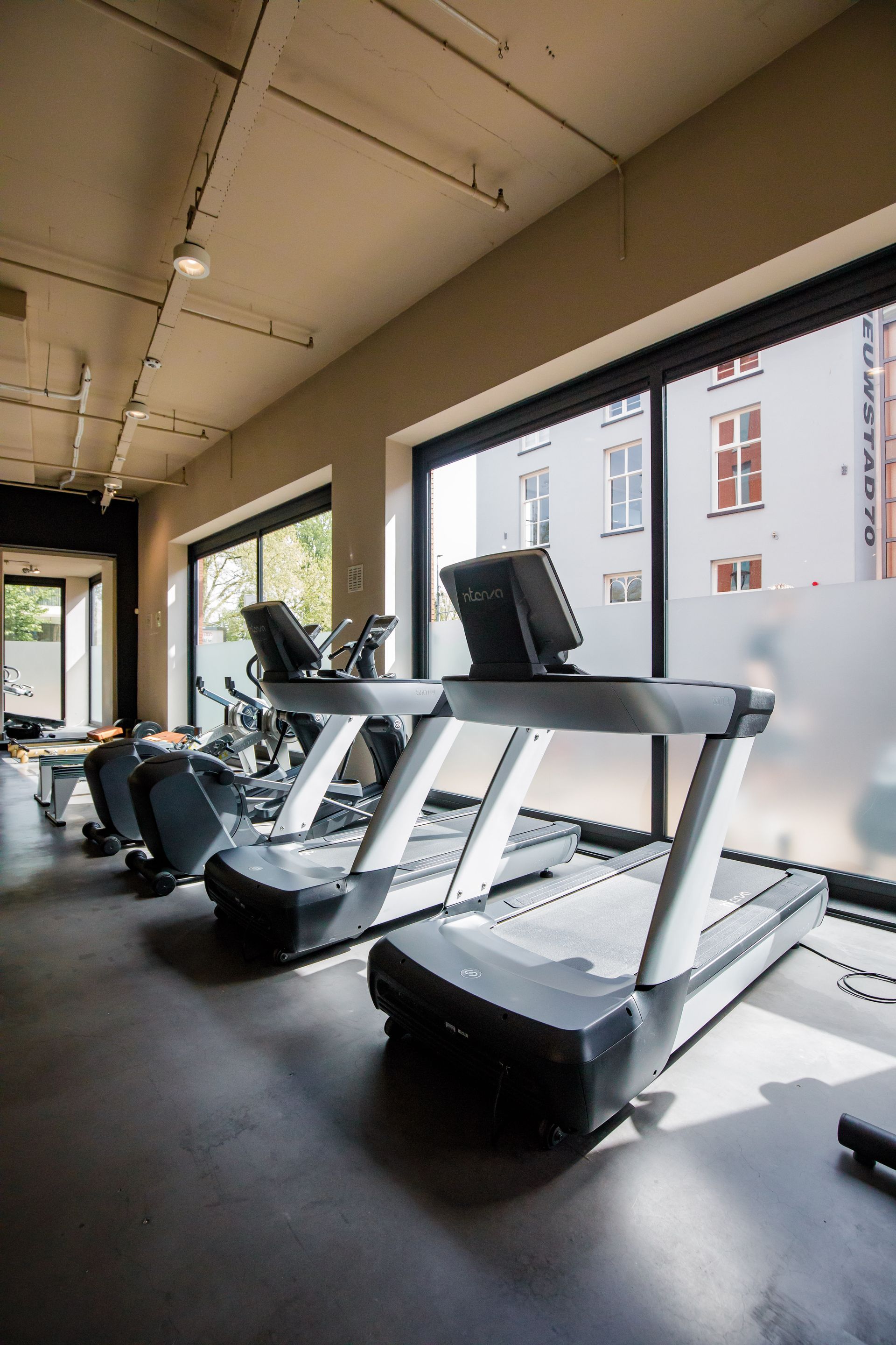 Een rij loopbanden in een sportschool naast een raam.