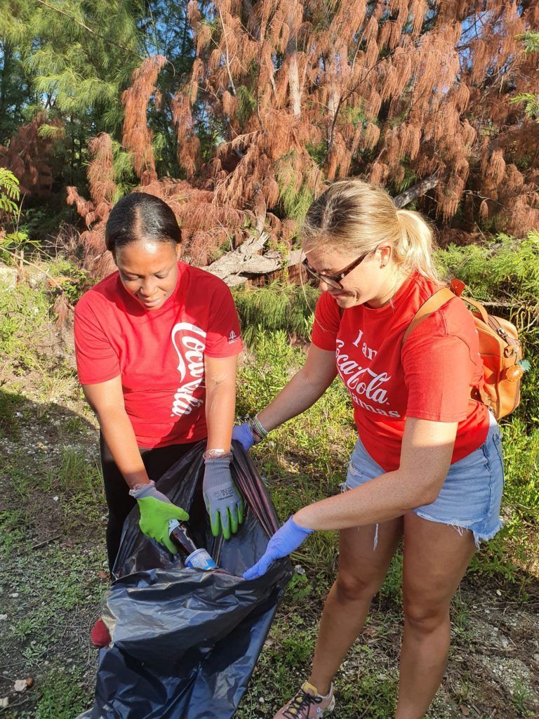 Two women in red Coca-Cola shirts cleaning up trash outdoors, one placing a bottle into a black trash bag.