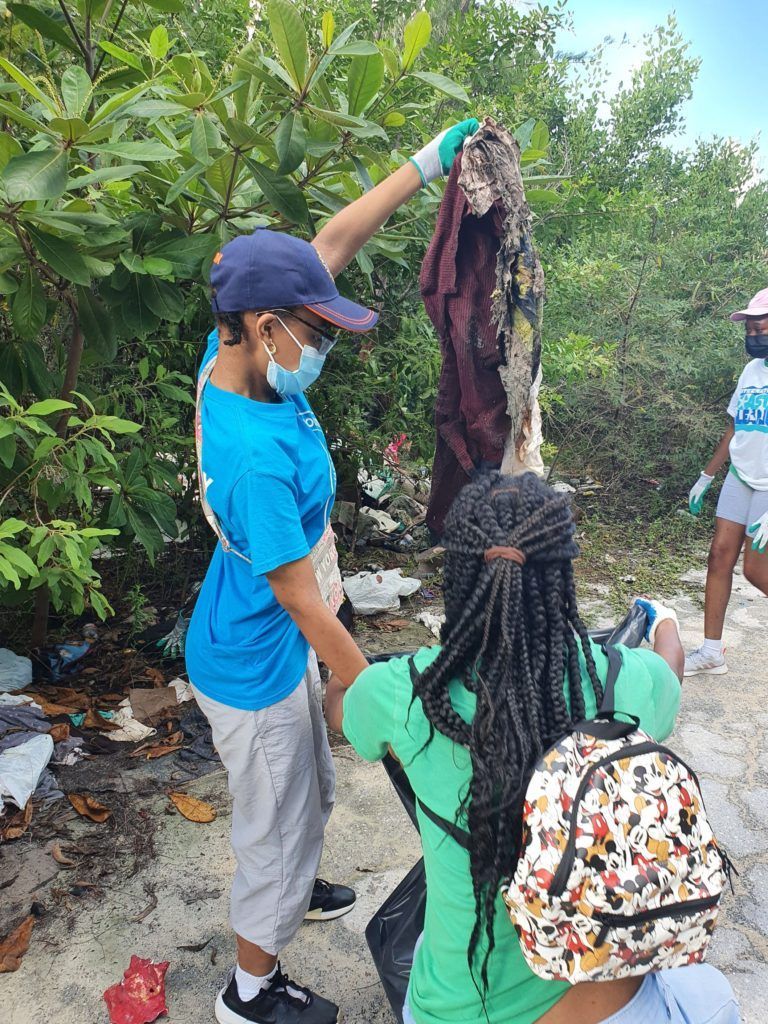 Volunteers in blue and green shirts clean up trash outdoors, one holding up a discarded garment. 