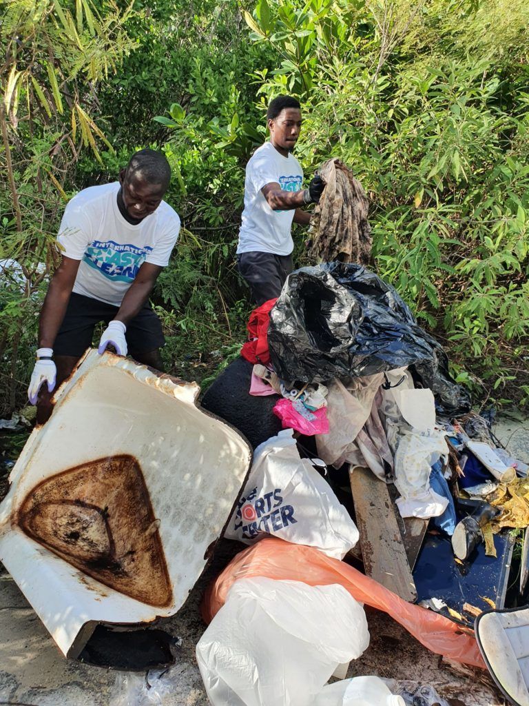 Two men in white shirts and gloves collect trash from a pile near green foliage;