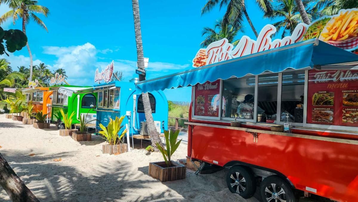 Colorful food trucks parked on a sandy beach under a clear blue sky. Palm trees and foliage frame the scene.
