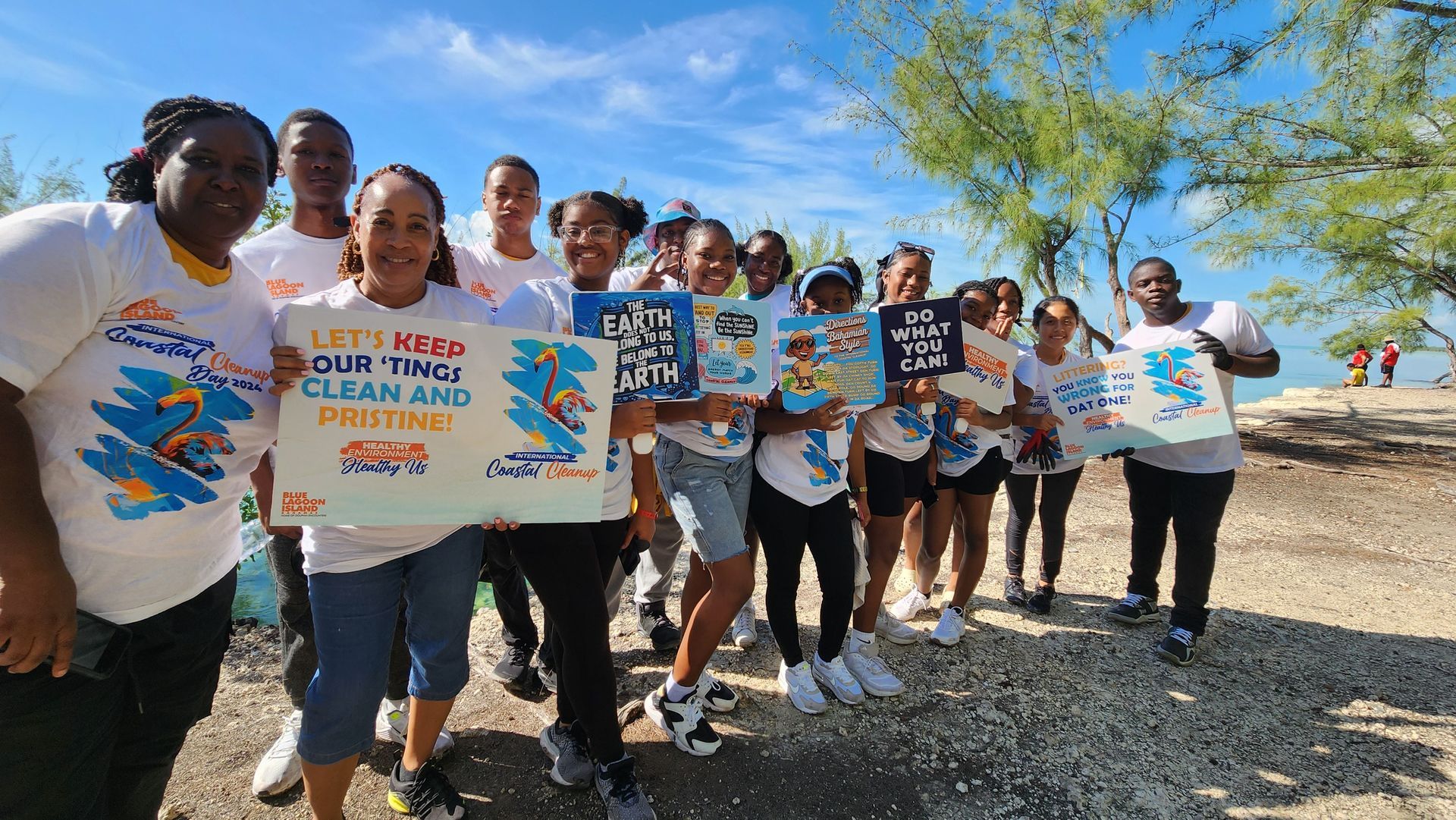 A large group of students in green shirts pose with black trash bags and a vehicle near a shoreline, smiling.
