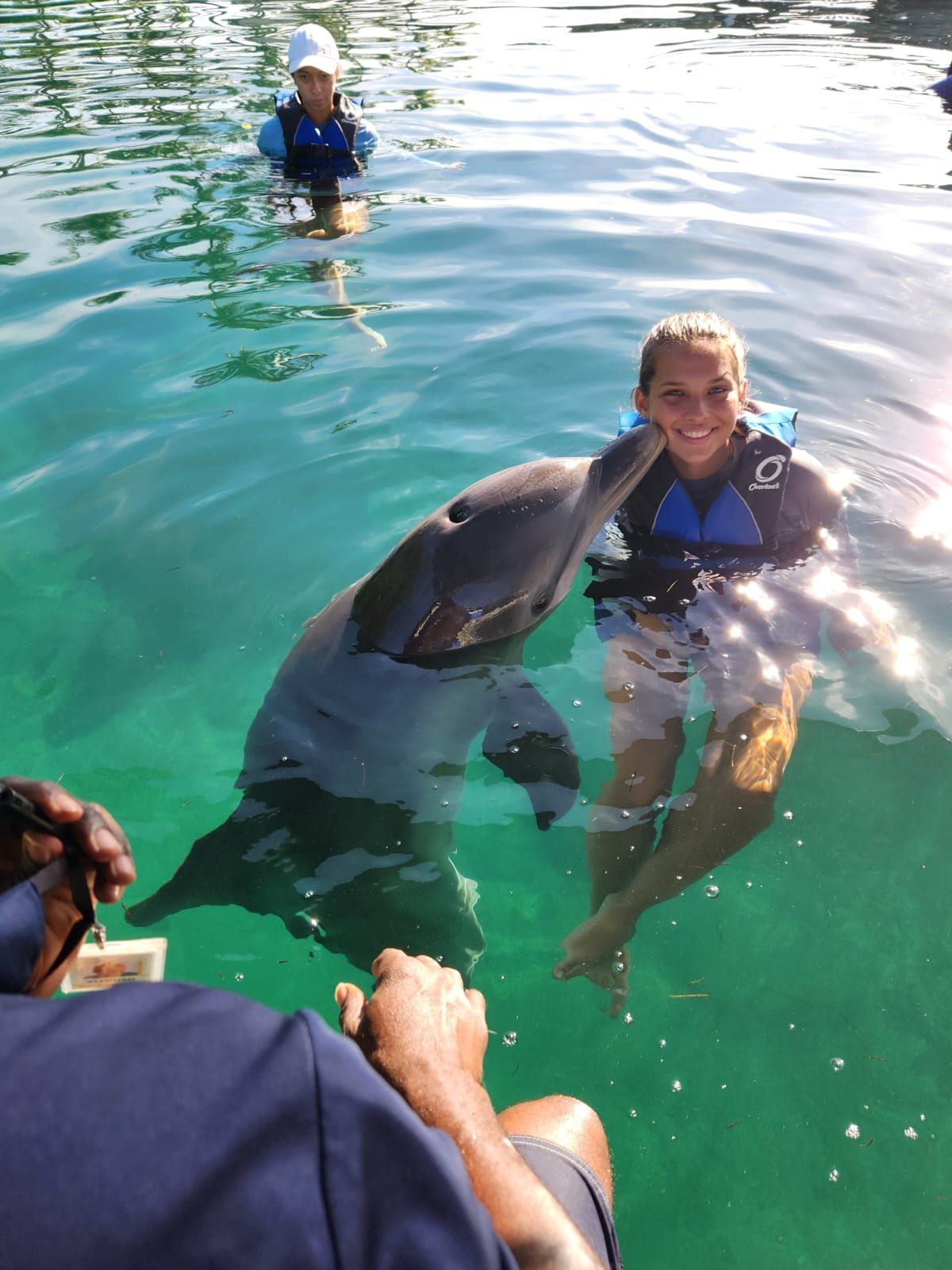 Woman in water with dolphin; dolphin touches her face. Another person in the water in the background.