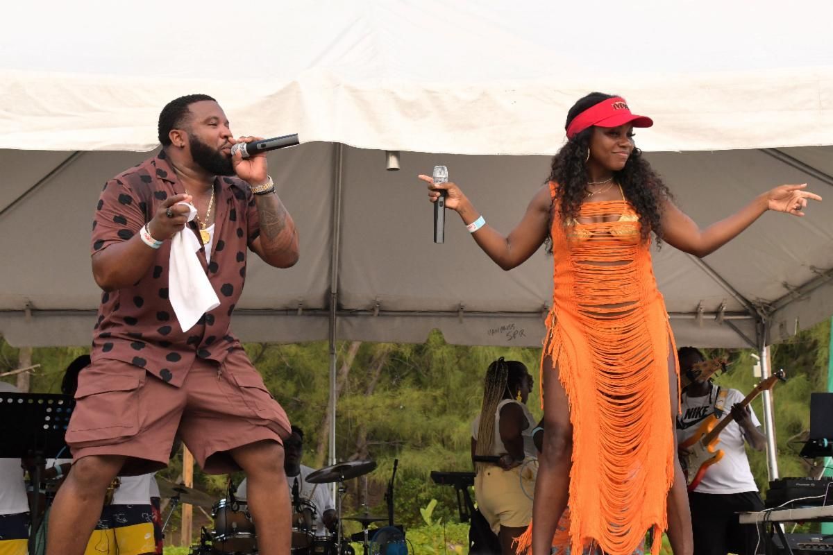 Two performers on stage at an outdoor event. A man sings with a microphone, and a woman in an orange dress dances.