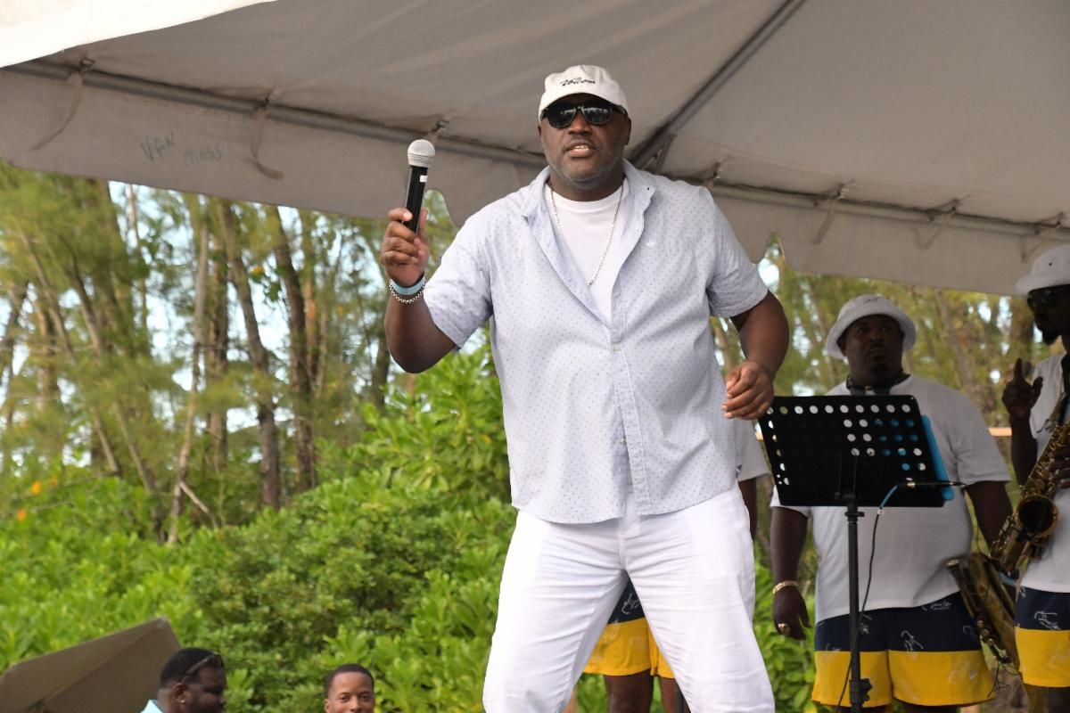 Man with microphone on stage, wearing white, gesturing. Band members in background. Outdoor setting.