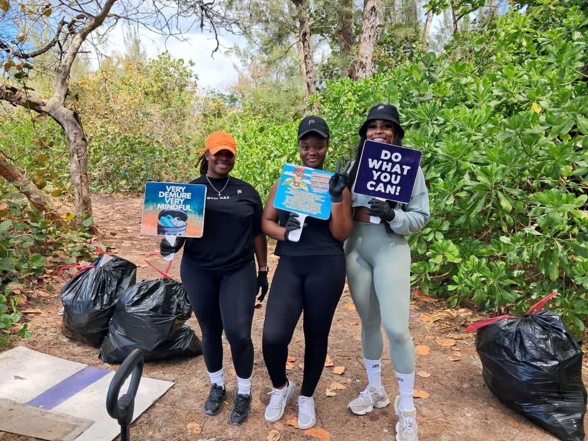Three people in athletic wear holding signs and garbage bags while volunteering outdoors, smiling at the camera.