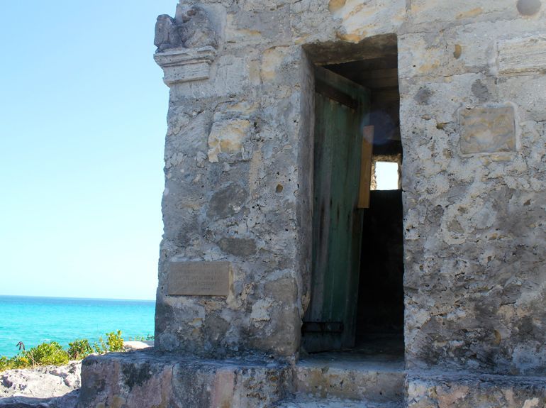 Stone structure with a turquoise ocean view. A green door is ajar, revealing a dark interior.
