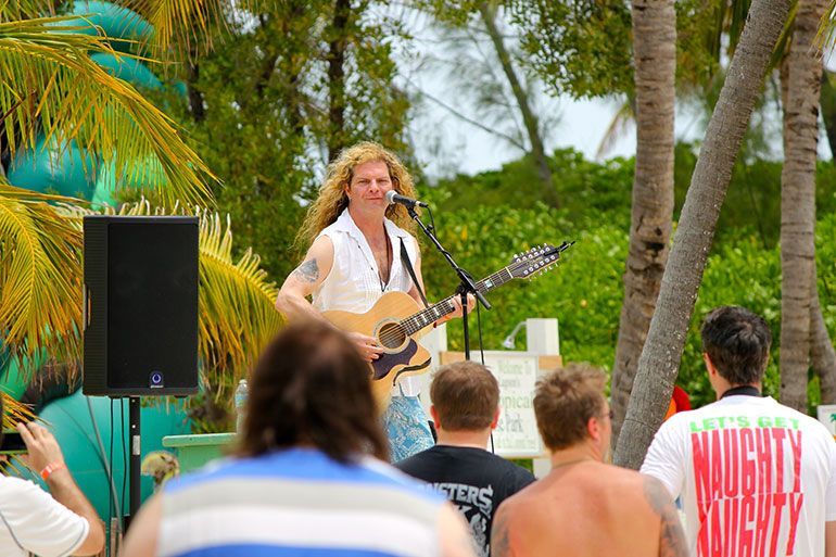 Man with long blond hair singing and playing guitar outdoors for an audience.