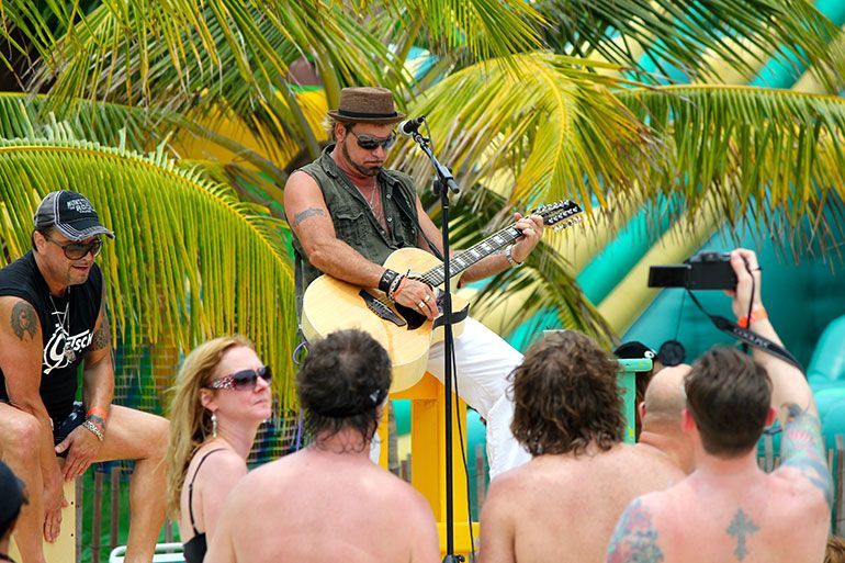 Musician with a hat and guitar performs on a stage near a pool, surrounded by an audience with a sunny backdrop.