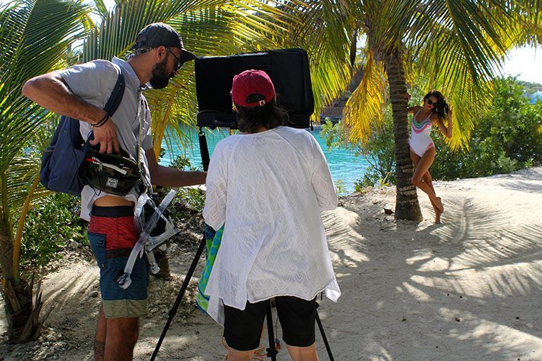 Two people filming a woman posing in a white swimsuit on a beach, with palms and ocean in the background.