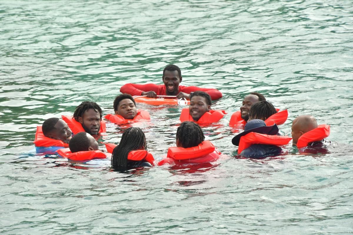 People in orange life vests in a circle in water; one man holds an orange float and smiles.