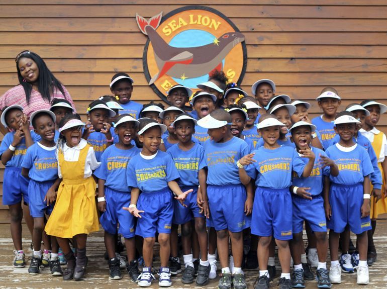 A group of children in blue shirts and shorts with a woman smile at the camera in front of a Sea Lion exhibit sign.