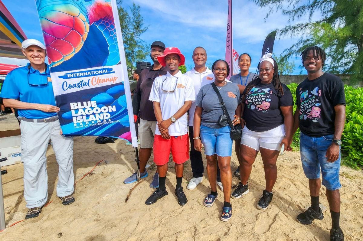 Group of people on a beach with a sign that says “Coastal Cleanup.” People smiling, sunny day.