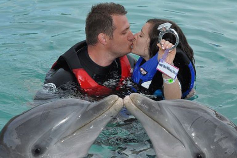 Couple kissing with two dolphins, holding a ring, in water.