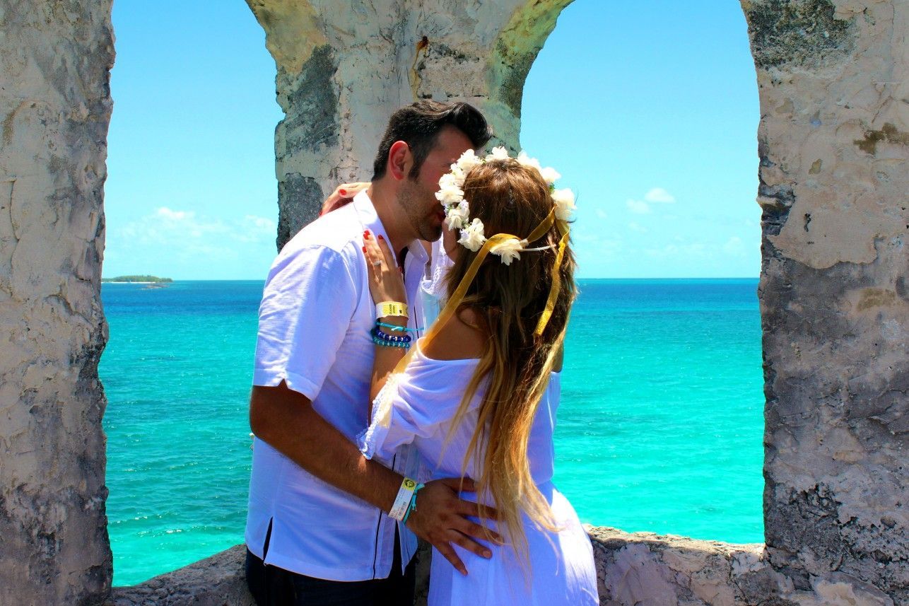 Couple kissing in a stone archway overlooking turquoise ocean; woman wears white dress and flower crown.