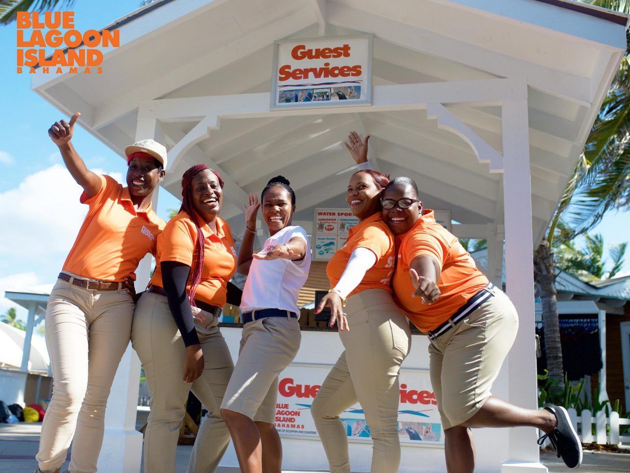 Five smiling women in orange shirts and khaki pants pose in front of a 