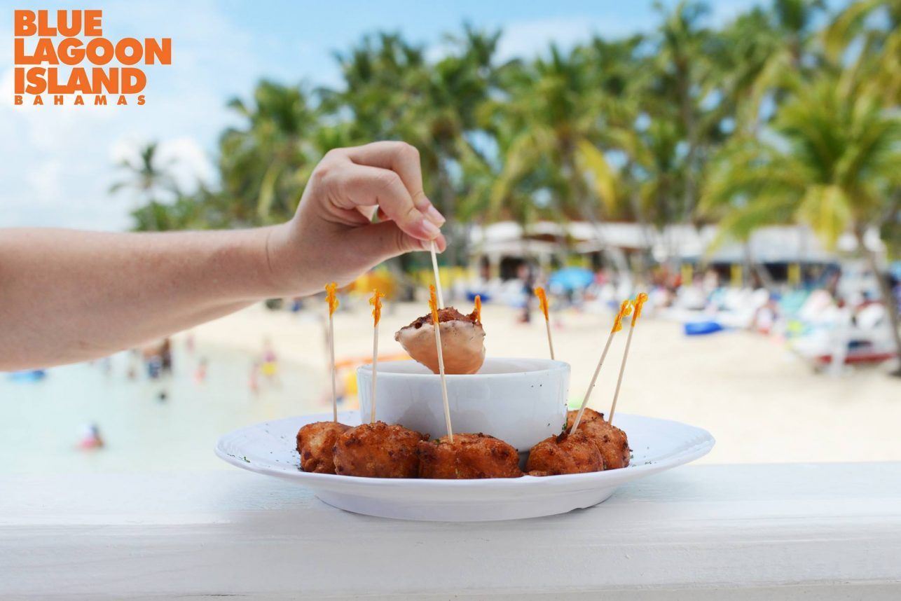 Hand reaching for fried food on a white plate at Blue Lagoon Island, Bahamas. Beach and palm trees in the blurred background.