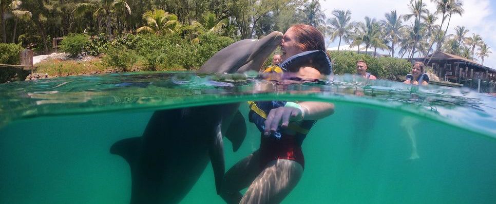 A woman in a life vest smiling as a dolphin kisses her cheek in clear turquoise water.