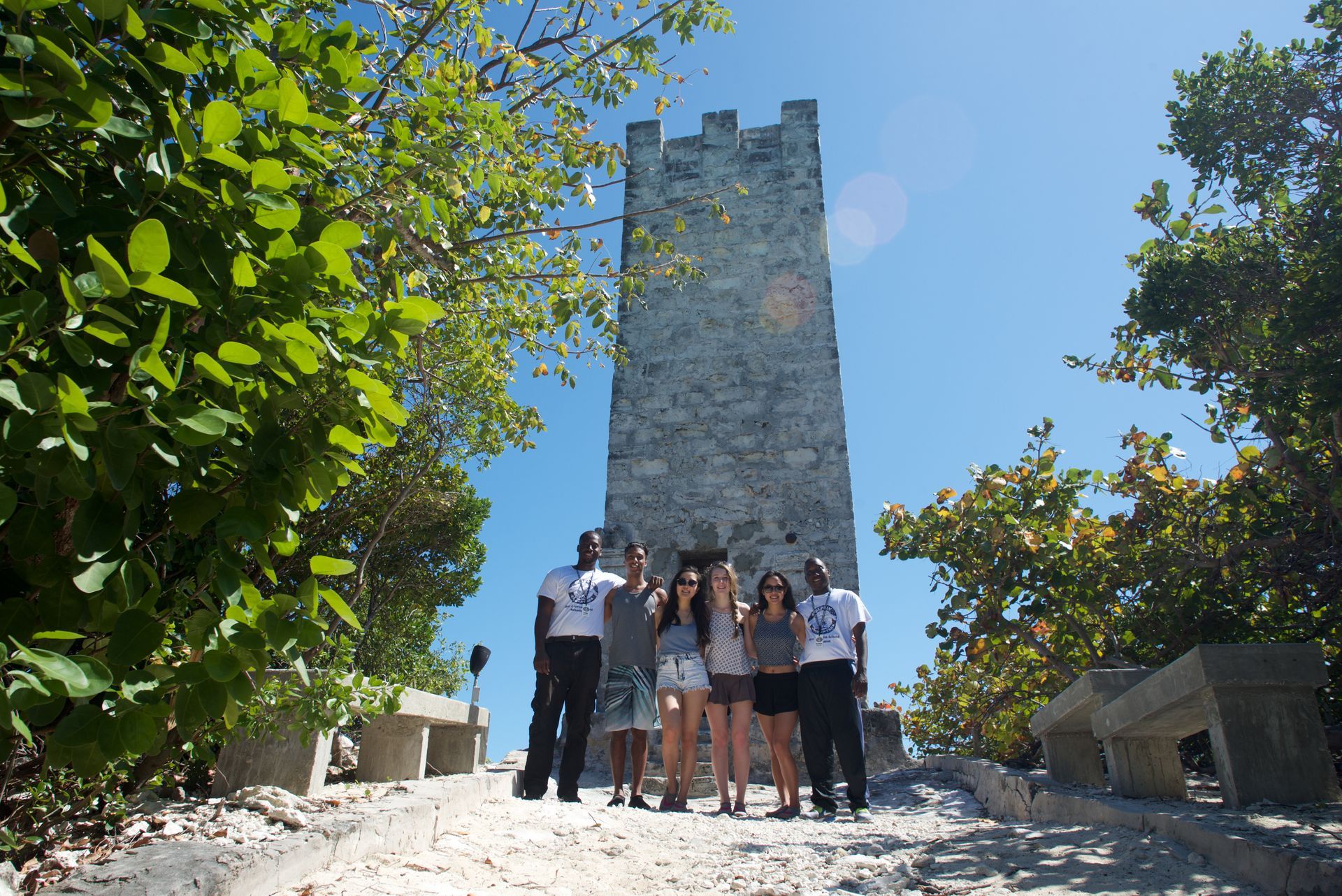 A group of people pose in front of a stone tower under a clear blue sky. Green foliage frames the shot.