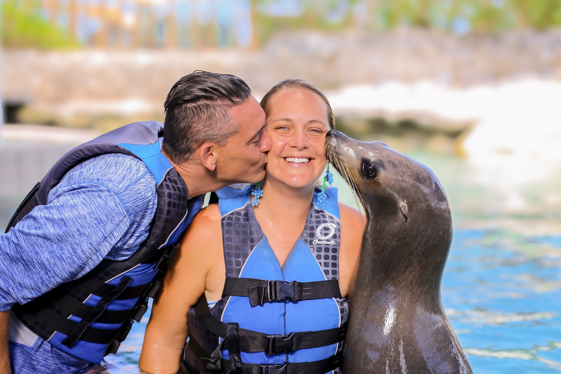 A woman smiles as a sea lion and a man kiss her cheeks, all wearing blue life vests, in the water.