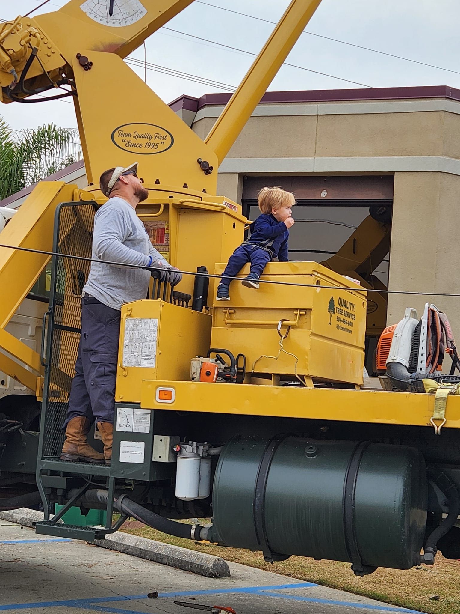 A person and a toddler sit on a large, yellow utility truck with a crane attachment, both looking upward.