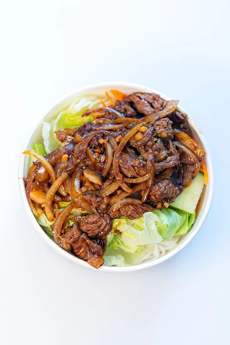 Sauté de bœuf et d'oignons sur un lit de laitue et de nouilles, servi dans un bol blanc sur fond blanc.