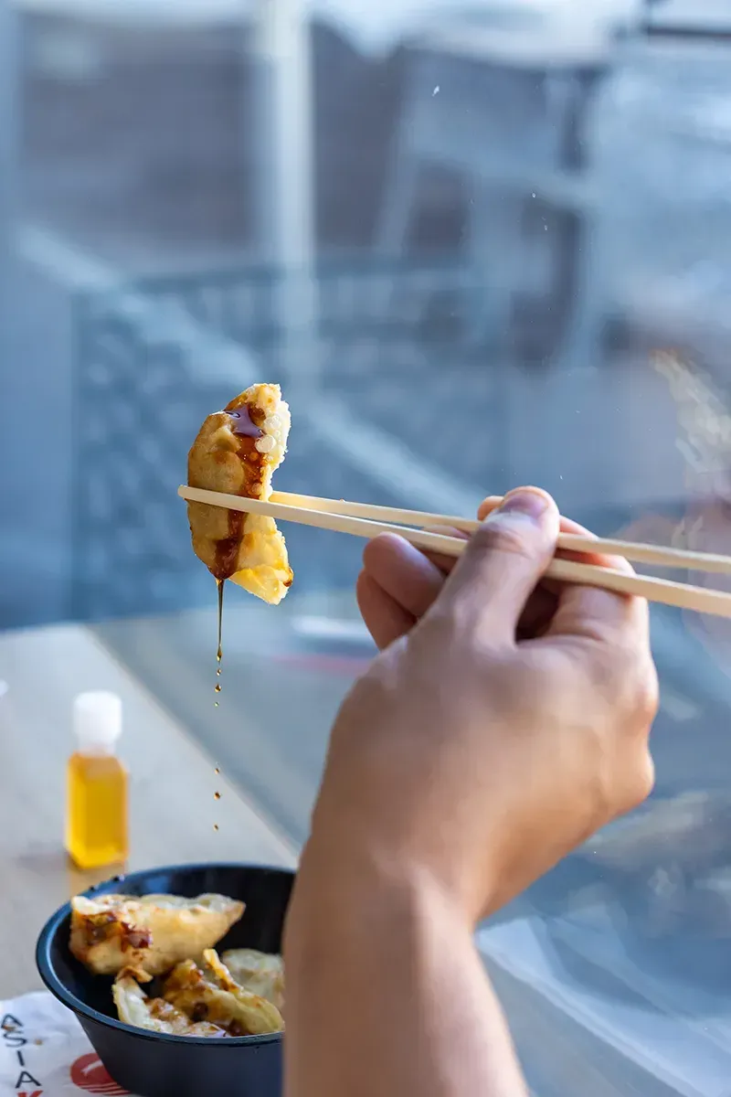 Une main tient un ravioli avec des baguettes et le trempe dans la sauce. Un bol de raviolis, une petite bouteille et une fenêtre sont visibles en arrière-plan.
