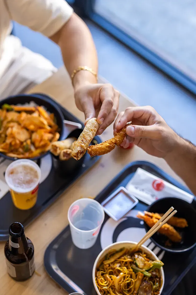 Des mains partageant des aliments frits à une table avec des plats asiatiques, des boissons et une fenêtre.