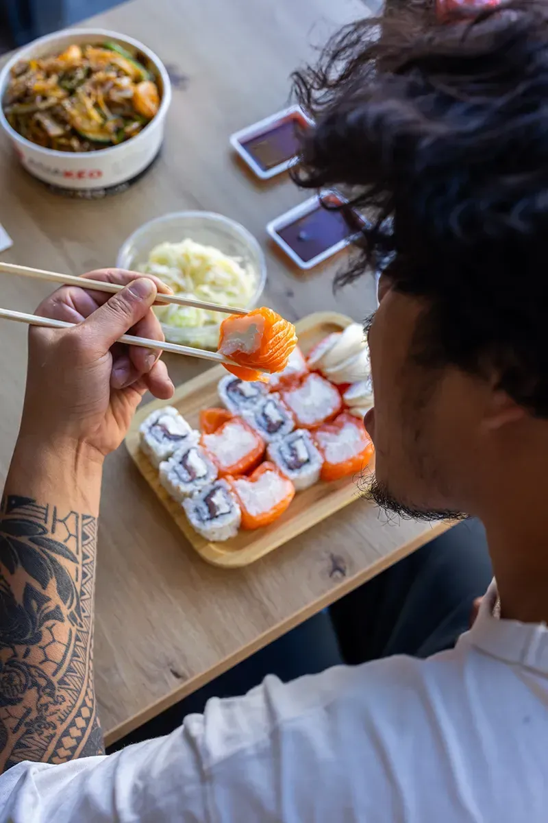 Une personne mange des sushis avec des baguettes ; table en bois, contenants à emporter et sauce soja.