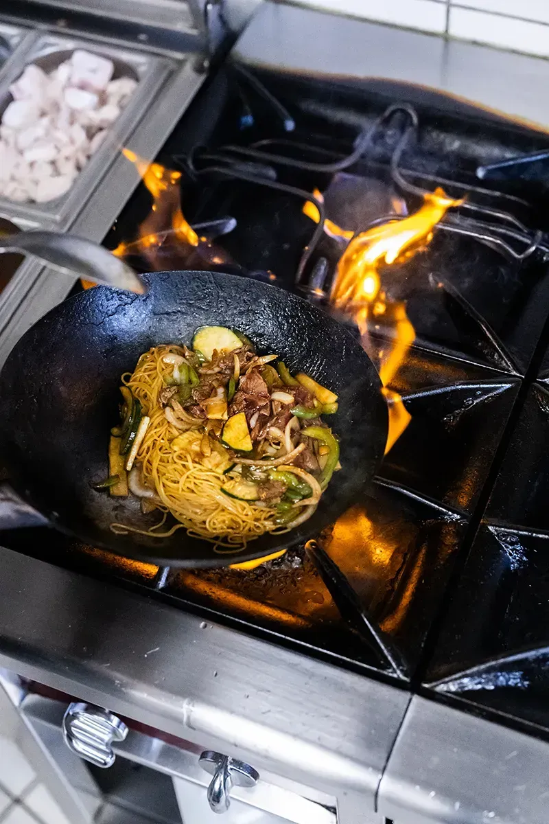 Wok avec des flammes pour cuire des nouilles, des légumes et de la viande sur une cuisinière professionnelle.