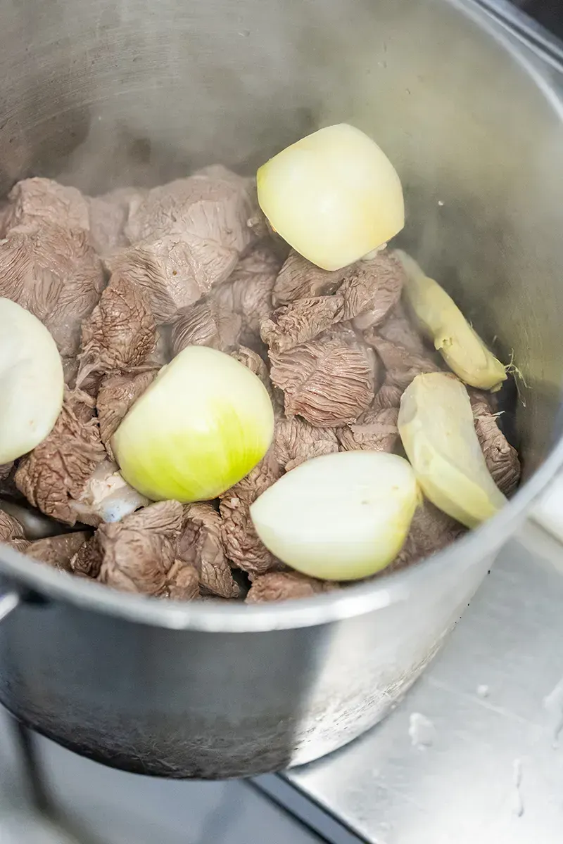 Des morceaux de bœuf, des demi-oignons et du gingembre mijotent dans une casserole en acier inoxydable. De la vapeur s'en échappe.