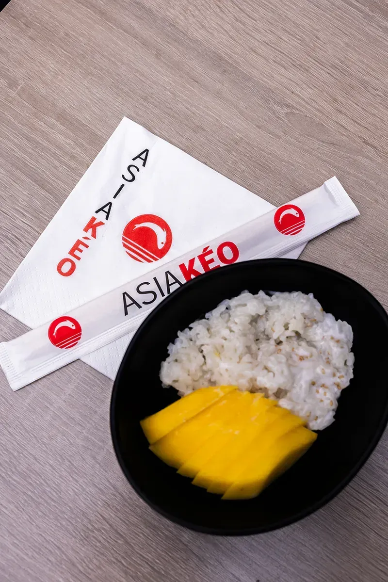Riz gluant à la mangue dans un bol noir avec baguettes et serviette, logo « ASIAKÉO ».