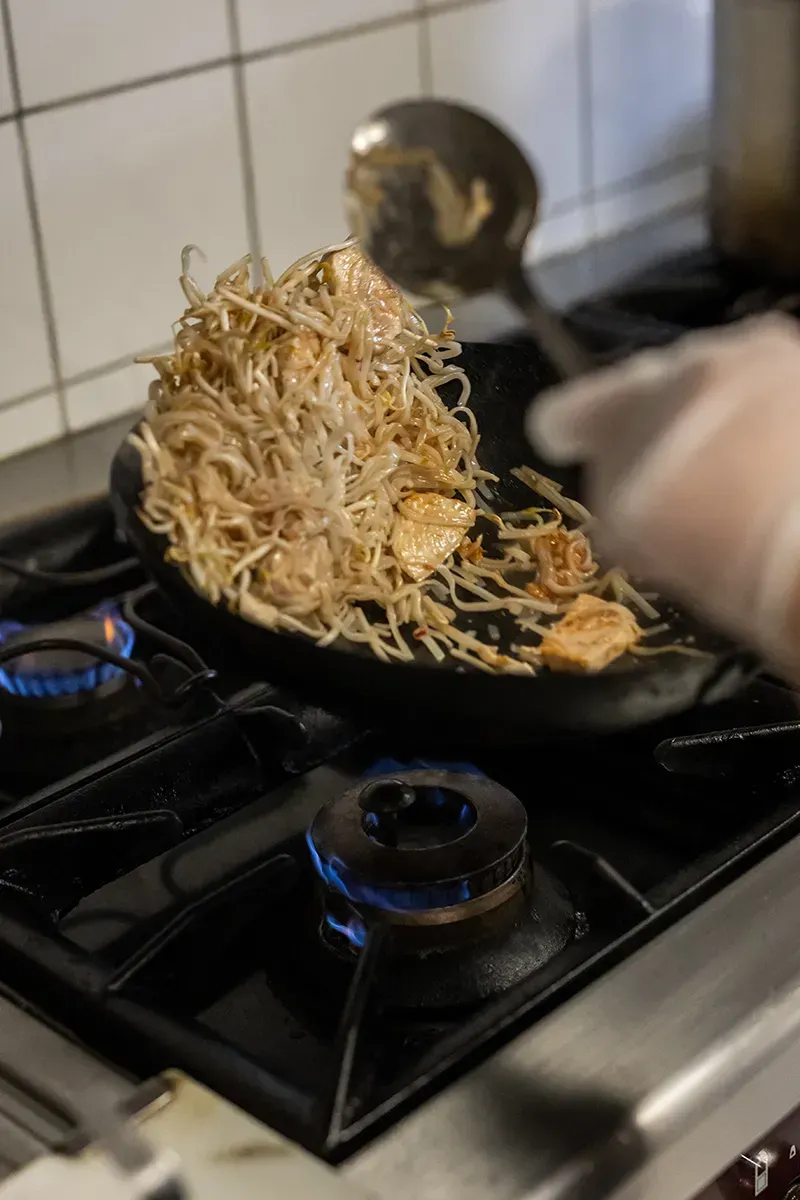 On fait sauter des aliments dans une poêle sur une cuisinière à gaz. Une main gantée tient une louche et mélange les ingrédients.