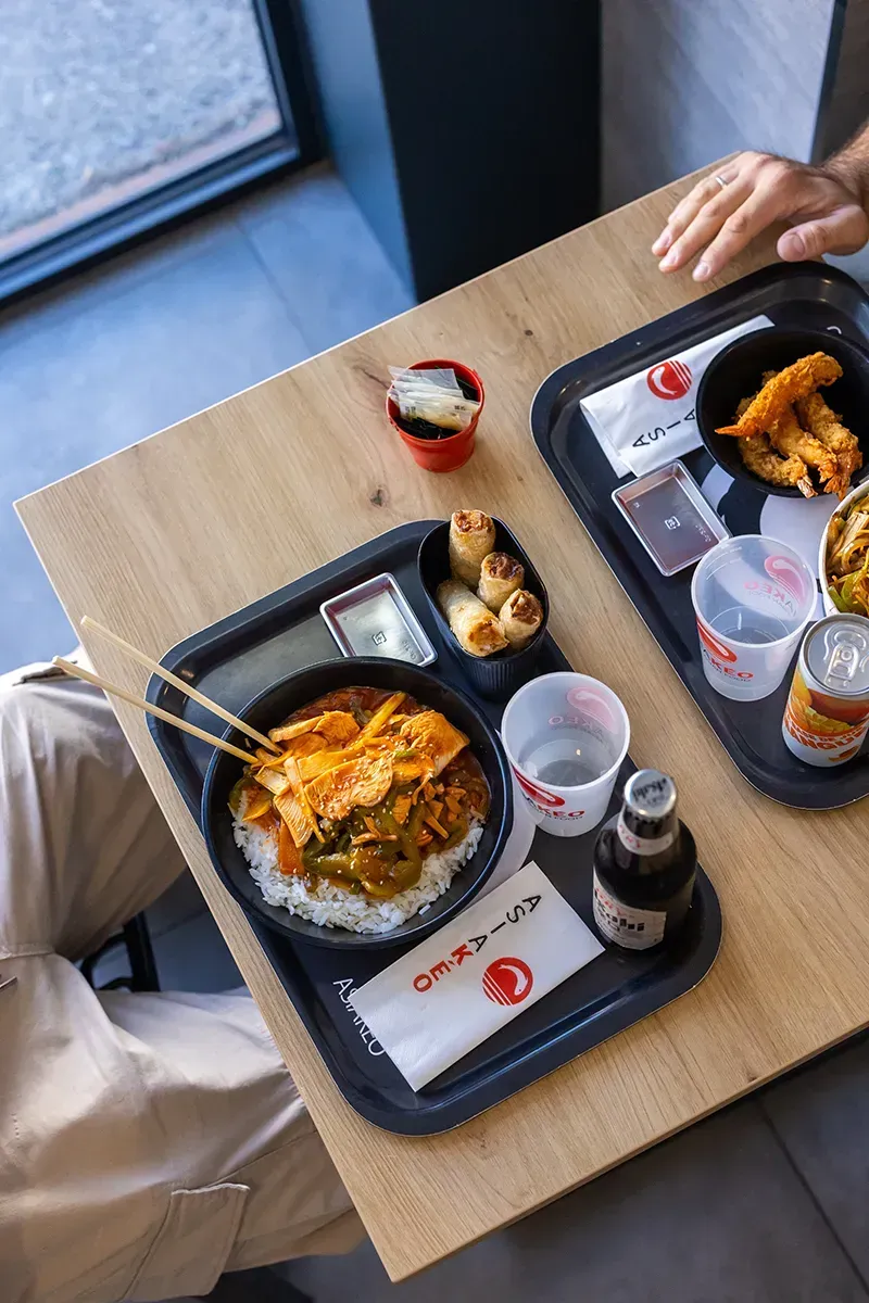 Deux plateaux noirs de nourriture asiatique sont posés sur une table en bois. On aperçoit une personne qui mange. Le menu comprend des bols de riz, des rouleaux de printemps et des boissons.