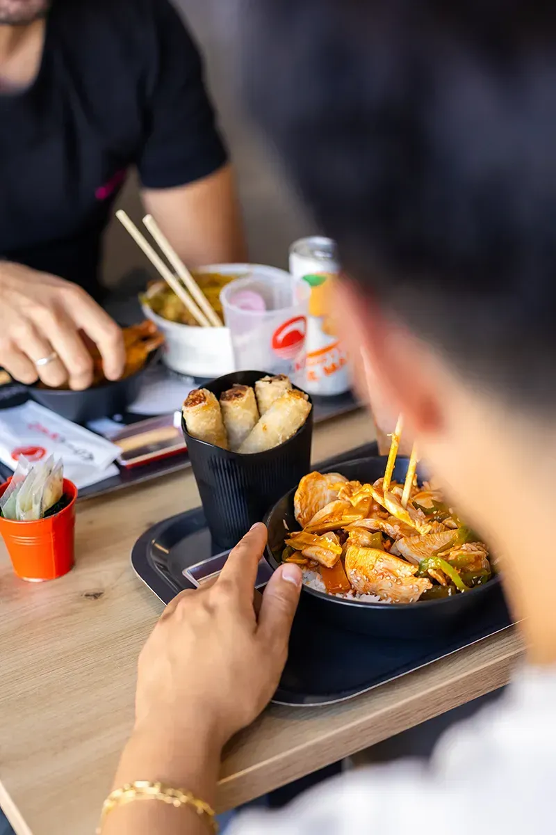 Des personnes mangent des plats asiatiques à une table. On aperçoit des rouleaux de printemps, des nouilles et des boissons.
