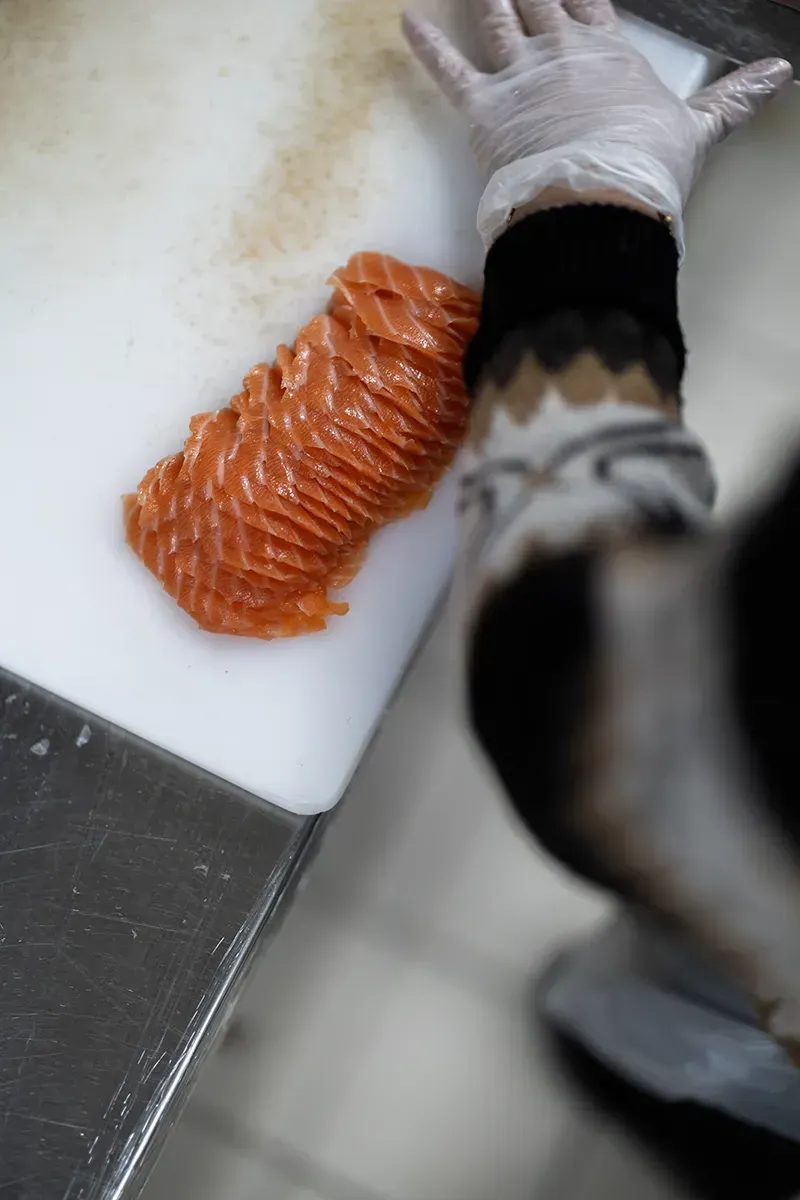 Des tranches de saumon cru sur une planche à découper blanche, à côté d'une main gantée dans une manche à motifs.