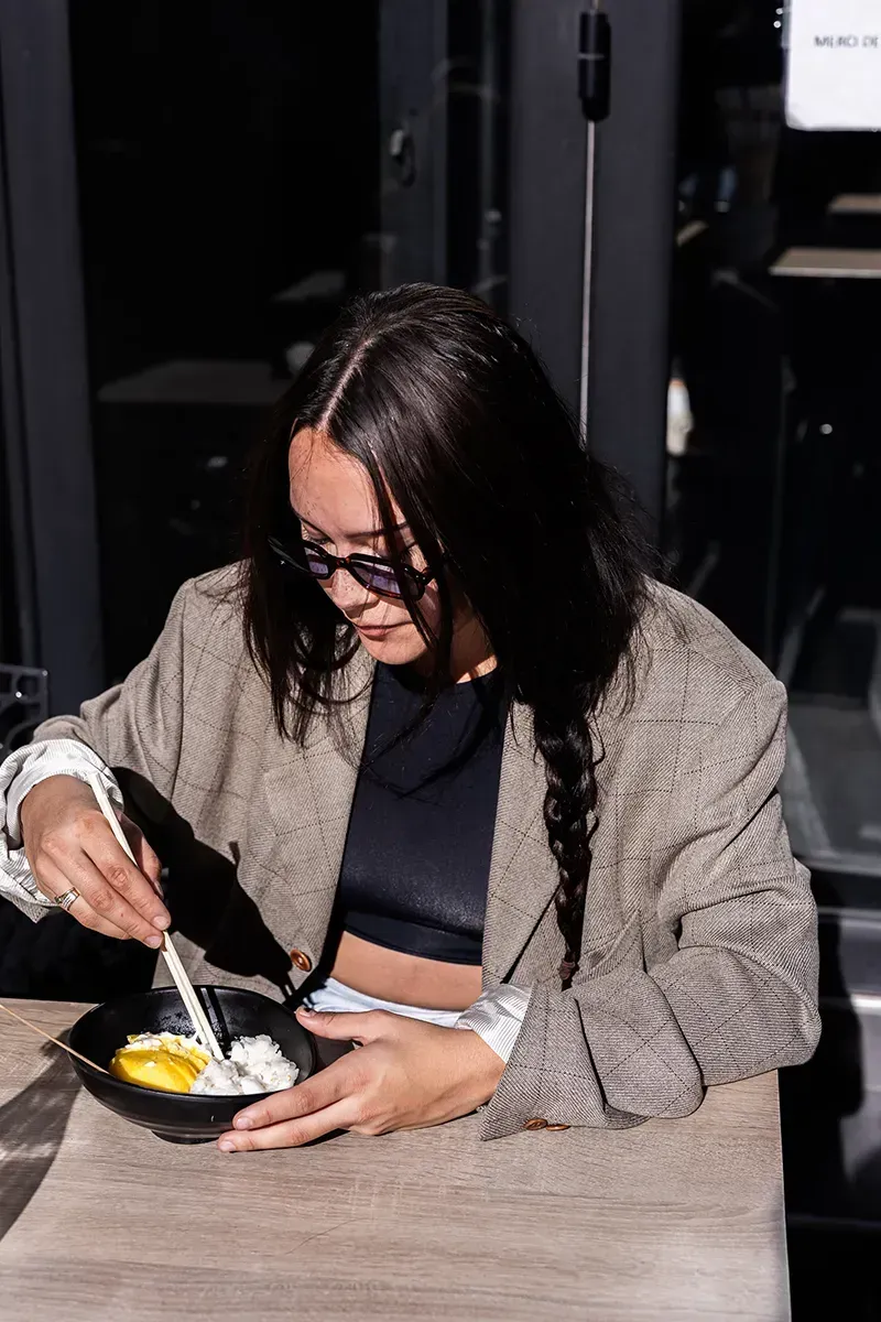 Une personne mangeant dans un bol avec des baguettes, portant des lunettes et un blazer, assise à une table.