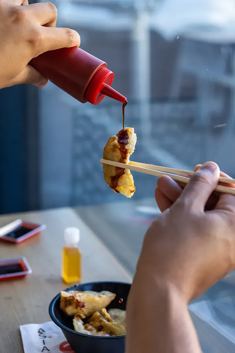 Une personne utilise des baguettes pour tenir sa nourriture tout en y versant de la sauce provenant d'une bouteille rouge.