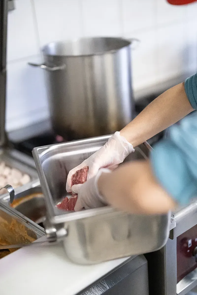 Des mains gantées manipulent de la viande crue dans un récipient en acier inoxydable, à côté d'un fourneau.