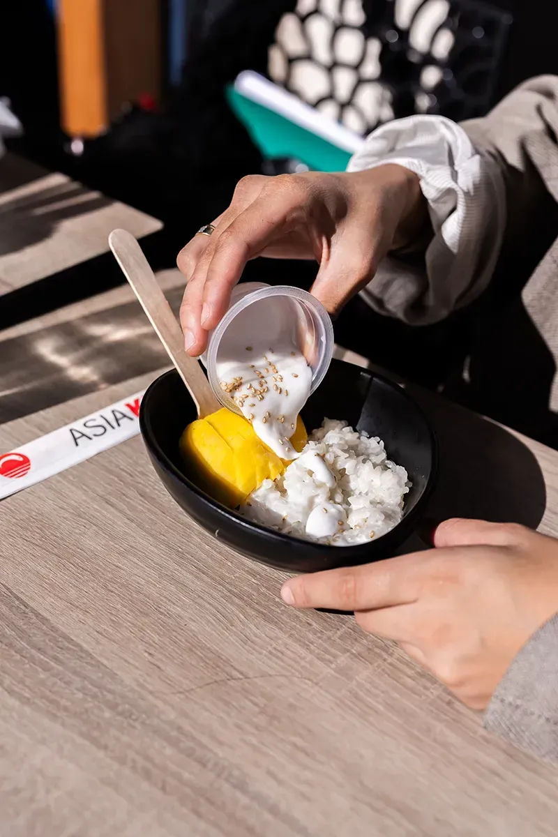 Riz gluant à la mangue arrosé de lait de coco dans un bol noir. Baguettes visibles, table en bois.