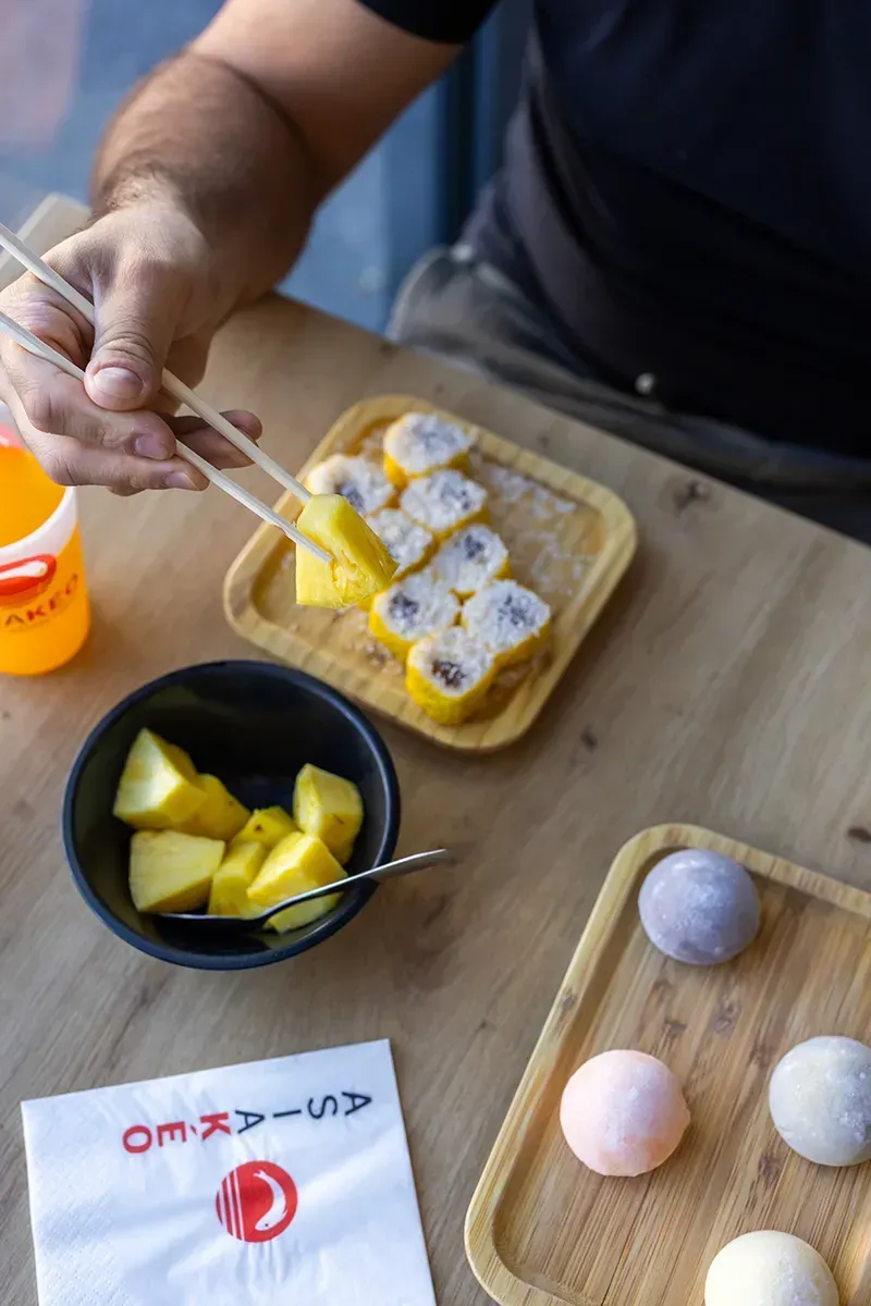 Une personne déguste des mochis avec des baguettes. Des tranches de mangue et des mochis sont présentés sur une table en bois.