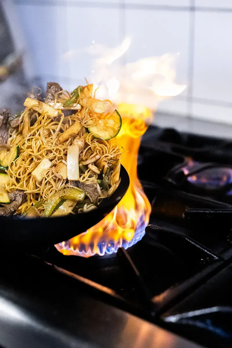 Wok contenant des nouilles et des légumes cuits dans les flammes sur une cuisinière à gaz.