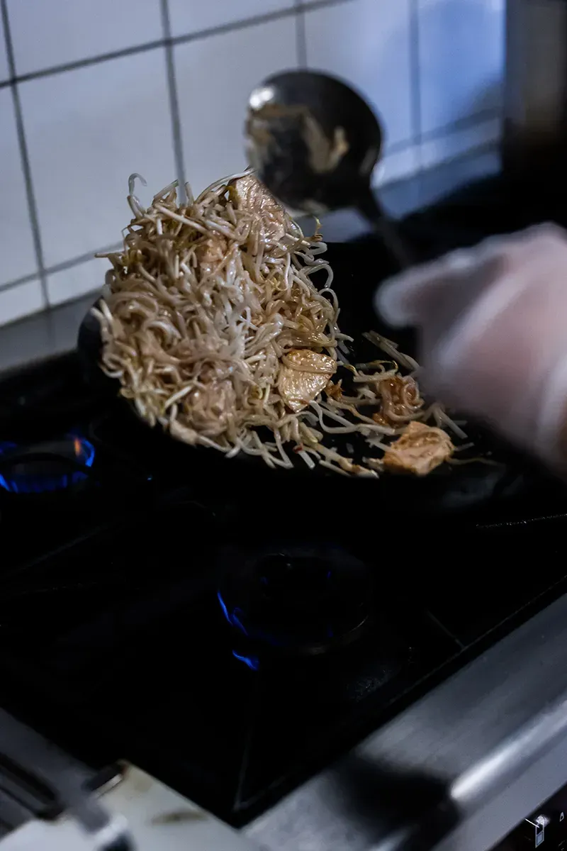 Une personne fait cuire des aliments sur une plaque de cuisson à l'aide d'une louche. Les aliments sont brun clair et emmêlés.